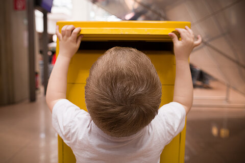 Ein Kind mit blonden kurzen Haaren ist von hinten zu sehen. Es trägt ein weißes Tshirt. Es steht vor einem gelben Briefkasten, in den es mit zwei Händen einen Brief einschmeißt.