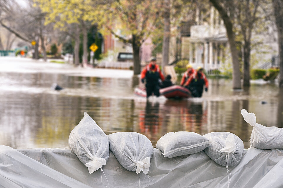 Eine Mauer mit Plastikfolie und Sandsäcken bedeckt. Im Hintergrund ein überflutete Straße und Häuser, ein Schlauchboot wird von Menschen durch das Wasser gezogen.