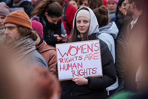 Eine Person steht in einer Menschenmenge. Sie guckt direkt in die Kamera und hält ein Poster mit der Aufschrift „Women’s Rights are Human Rights“ in der Hand.